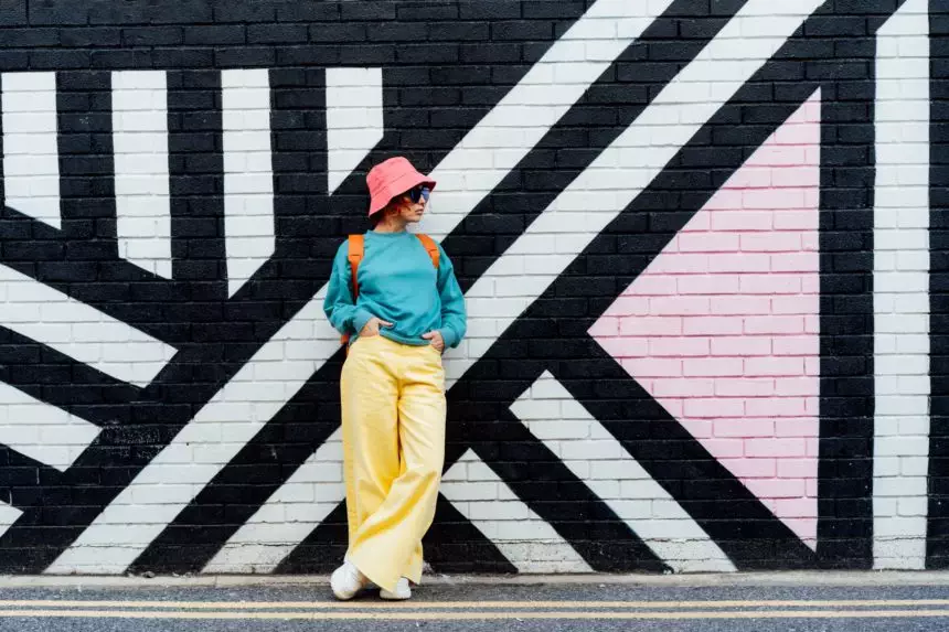 Hipster fashion woman in bright clothes, sun glasses and bucket hat posing on painted brick wall